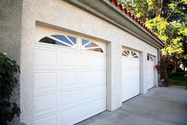 white garage door with white cemented wall, red bricked roof, green leaves on trees -repair garage doors